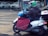 A person wearing a colorful helmet and dark jacket is seated on a white scooter parked at a crosswalk. The scooter is equipped with a large red cover, and there is a blue plastic bag on the footrest. Vehicles are visible in the background, along with a graffiti-covered sign.