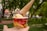 A hand holds a cup of ice cream featuring two scoops, one yellow and one purple, with an upside-down ice cream cone placed on top. The cup has vibrant red and yellow colors with 'Ice Cream' written on it, and there are blurred trees and a path in the background.