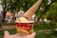 A hand holds a cup of ice cream featuring two scoops, one yellow and one purple, with an upside-down ice cream cone placed on top. The cup has vibrant red and yellow colors with 'Ice Cream' written on it, and there are blurred trees and a path in the background.