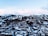 A scenic winter landscape featuring a snow-covered mountain range with ski trails visible. At the base of the mountains, a town with numerous snow-laden buildings is nestled among dense clusters of trees.