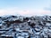 A scenic winter landscape featuring a snow-covered mountain range with ski trails visible. At the base of the mountains, a town with numerous snow-laden buildings is nestled among dense clusters of trees.