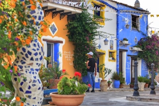 A vibrant street scene features colorful houses with bright blue and orange facades. The walls are adorned with decorative tiles and plants. A person wearing casual clothing and a white cap stands in front of the house, while various potted plants and colorful flowers embellish the scene. Intricate architectural details and outdoor decor create a lively, picturesque atmosphere.