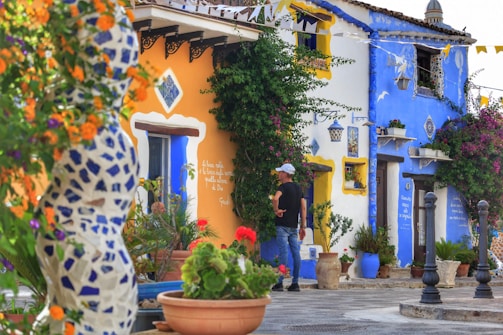 A vibrant street scene features colorful houses with bright blue and orange facades. The walls are adorned with decorative tiles and plants. A person wearing casual clothing and a white cap stands in front of the house, while various potted plants and colorful flowers embellish the scene. Intricate architectural details and outdoor decor create a lively, picturesque atmosphere.
