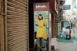 A street scene with a poster featuring a smiling woman in traditional Indian attire placed outside a shop. The poster has a vibrant background, and adjacent to it, various shop signs advertise different cuisines and services. The street is lined with shops and a few people can be seen walking in the distance.