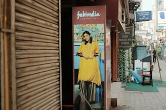 A street scene with a poster featuring a smiling woman in traditional Indian attire placed outside a shop. The poster has a vibrant background, and adjacent to it, various shop signs advertise different cuisines and services. The street is lined with shops and a few people can be seen walking in the distance.