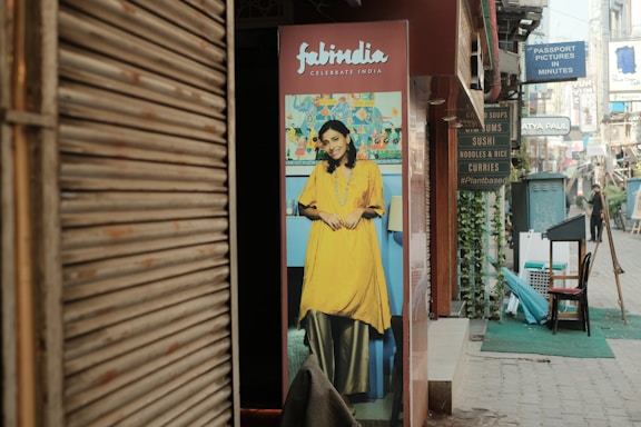 A street scene with a poster featuring a smiling woman in traditional Indian attire placed outside a shop. The poster has a vibrant background, and adjacent to it, various shop signs advertise different cuisines and services. The street is lined with shops and a few people can be seen walking in the distance.