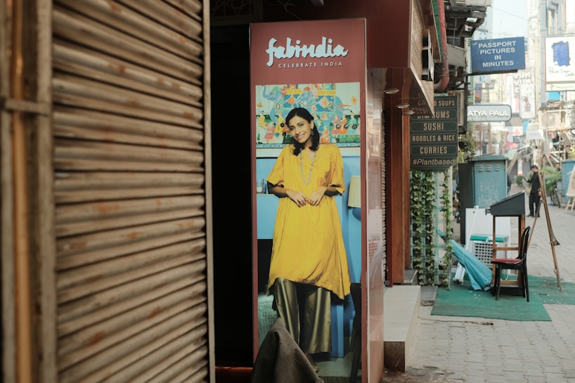 A street scene with a poster featuring a smiling woman in traditional Indian attire placed outside a shop. The poster has a vibrant background, and adjacent to it, various shop signs advertise different cuisines and services. The street is lined with shops and a few people can be seen walking in the distance.