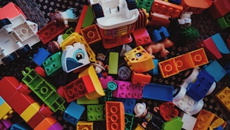A scattered collection of colorful building blocks and toy pieces, featuring various shapes and sizes. Some blocks have illustrations of animals and are interspersed with small toy figures. The toys are placed on a textured dark surface.