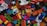 A scattered collection of colorful building blocks and toy pieces, featuring various shapes and sizes. Some blocks have illustrations of animals and are interspersed with small toy figures. The toys are placed on a textured dark surface.