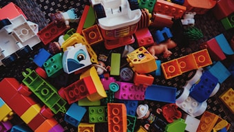 A scattered collection of colorful building blocks and toy pieces, featuring various shapes and sizes. Some blocks have illustrations of animals and are interspersed with small toy figures. The toys are placed on a textured dark surface.
