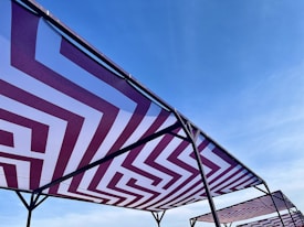 Geometric-patterned canopies with maroon and white zigzag designs are stretched over metal frames against a clear blue sky.