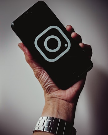 A hand holding a smartphone with a dark screen displaying a prominent Instagram logo. The hand is wearing a silver metallic watch, and the background is softly lit, creating a subtle shadow effect.