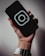 A hand holding a smartphone with a dark screen displaying a prominent Instagram logo. The hand is wearing a silver metallic watch, and the background is softly lit, creating a subtle shadow effect.