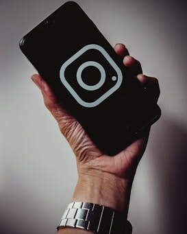 A hand holding a smartphone with a dark screen displaying a prominent Instagram logo. The hand is wearing a silver metallic watch, and the background is softly lit, creating a subtle shadow effect.