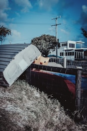 A metallic structure or part of a vehicle is seen leaning against a wire fence, set on a grassy patch. In the background, there is a modern-looking building with large windows, a tree, and a telephone pole with wires stretching across a blue sky.