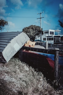 A metallic structure or part of a vehicle is seen leaning against a wire fence, set on a grassy patch. In the background, there is a modern-looking building with large windows, a tree, and a telephone pole with wires stretching across a blue sky.