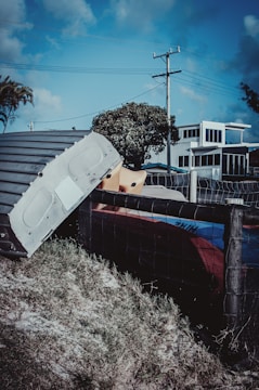 A metallic structure or part of a vehicle is seen leaning against a wire fence, set on a grassy patch. In the background, there is a modern-looking building with large windows, a tree, and a telephone pole with wires stretching across a blue sky.