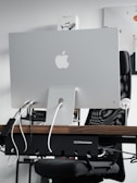 A sleek modern workspace featuring the back of an Apple iMac computer, with several cables plugged into it. The computer is positioned on a wooden desk, with a mesh office chair behind it. A product box labeled 'Mavic Air 2' is visible on the shelf above the desk. The atmosphere is minimalistic and tech-oriented.