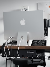 A sleek modern workspace featuring the back of an Apple iMac computer, with several cables plugged into it. The computer is positioned on a wooden desk, with a mesh office chair behind it. A product box labeled 'Mavic Air 2' is visible on the shelf above the desk. The atmosphere is minimalistic and tech-oriented.