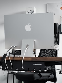 A sleek modern workspace featuring the back of an Apple iMac computer, with several cables plugged into it. The computer is positioned on a wooden desk, with a mesh office chair behind it. A product box labeled 'Mavic Air 2' is visible on the shelf above the desk. The atmosphere is minimalistic and tech-oriented.