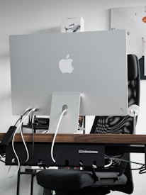 A sleek modern workspace featuring the back of an Apple iMac computer, with several cables plugged into it. The computer is positioned on a wooden desk, with a mesh office chair behind it. A product box labeled 'Mavic Air 2' is visible on the shelf above the desk. The atmosphere is minimalistic and tech-oriented.