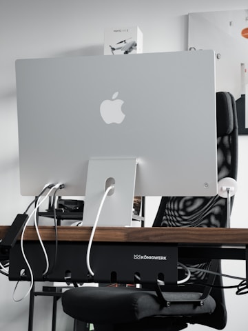 A sleek modern workspace featuring the back of an Apple iMac computer, with several cables plugged into it. The computer is positioned on a wooden desk, with a mesh office chair behind it. A product box labeled 'Mavic Air 2' is visible on the shelf above the desk. The atmosphere is minimalistic and tech-oriented.