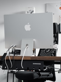 A sleek modern workspace featuring the back of an Apple iMac computer, with several cables plugged into it. The computer is positioned on a wooden desk, with a mesh office chair behind it. A product box labeled 'Mavic Air 2' is visible on the shelf above the desk. The atmosphere is minimalistic and tech-oriented.