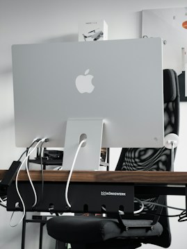 A sleek modern workspace featuring the back of an Apple iMac computer, with several cables plugged into it. The computer is positioned on a wooden desk, with a mesh office chair behind it. A product box labeled 'Mavic Air 2' is visible on the shelf above the desk. The atmosphere is minimalistic and tech-oriented.
