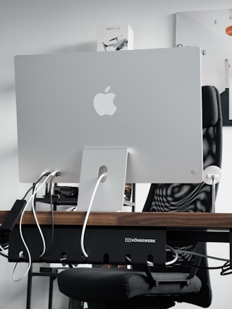A sleek modern workspace featuring the back of an Apple iMac computer, with several cables plugged into it. The computer is positioned on a wooden desk, with a mesh office chair behind it. A product box labeled 'Mavic Air 2' is visible on the shelf above the desk. The atmosphere is minimalistic and tech-oriented.