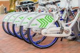 A row of neatly aligned white bicycles with green geometric patterns parked on a brick-paved area. Each bike has a blue wheel trim and is part of a bike-sharing system. They are locked in a docking station, indicating an urban, eco-friendly transportation option.