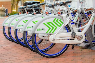 A row of neatly aligned white bicycles with green geometric patterns parked on a brick-paved area. Each bike has a blue wheel trim and is part of a bike-sharing system. They are locked in a docking station, indicating an urban, eco-friendly transportation option.