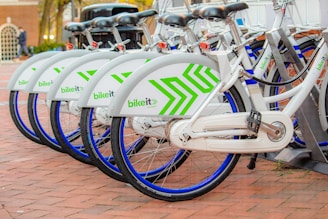 A row of neatly aligned white bicycles with green geometric patterns parked on a brick-paved area. Each bike has a blue wheel trim and is part of a bike-sharing system. They are locked in a docking station, indicating an urban, eco-friendly transportation option.