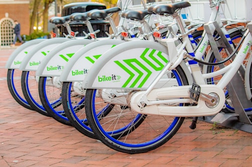 A row of neatly aligned white bicycles with green geometric patterns parked on a brick-paved area. Each bike has a blue wheel trim and is part of a bike-sharing system. They are locked in a docking station, indicating an urban, eco-friendly transportation option.