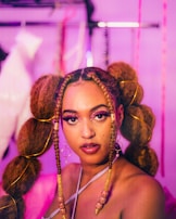 A woman is looking directly at the camera with a confident expression. Her hair is styled in large braids adorned with decorative elements and gold accessories. The makeup is bold, featuring bright eyeshadow and glitter. She wears large hoop earrings, and the background is blurred with shades of pink and purple.