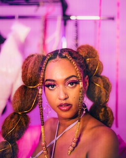 A woman is looking directly at the camera with a confident expression. Her hair is styled in large braids adorned with decorative elements and gold accessories. The makeup is bold, featuring bright eyeshadow and glitter. She wears large hoop earrings, and the background is blurred with shades of pink and purple.
