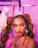 A woman is looking directly at the camera with a confident expression. Her hair is styled in large braids adorned with decorative elements and gold accessories. The makeup is bold, featuring bright eyeshadow and glitter. She wears large hoop earrings, and the background is blurred with shades of pink and purple.