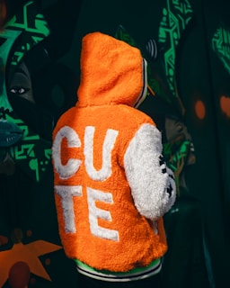 A person is wearing an orange and white fluffy jacket with the word 'CUTE' displayed in large white letters on the back. The background features dark green graffiti art with abstract shapes and a stylized face.