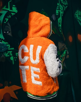 A person is wearing an orange and white fluffy jacket with the word 'CUTE' displayed in large white letters on the back. The background features dark green graffiti art with abstract shapes and a stylized face.