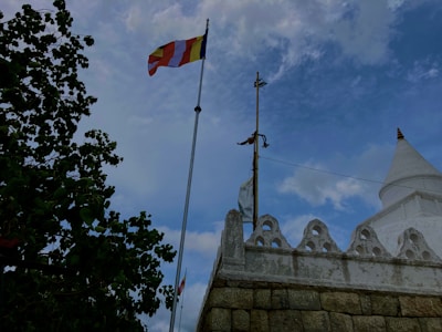 A flag with multiple colors is prominently displayed on a flagpole against a backdrop of a partly cloudy blue sky. To the right, there is a white stupa, partially visible, with traditional architectural design and a circular base. A large leafy tree occupies the left side of the image, providing contrast with its dark green leaves.