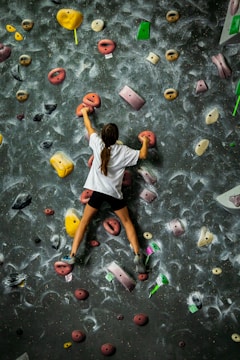 A person is climbing an indoor bouldering wall with various colorful holds. The wall has a rough surface and is covered in chalk marks, indicating frequent use. The climber is wearing a white shirt and black shorts, concentrating on moving upwards.