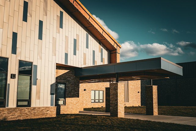 A modern building with a distinctive angular roof and light-colored paneling, featuring a covered entrance supported by brick pillars. The structure is set against a deep blue sky with scattered clouds, casting shadows on the walls.