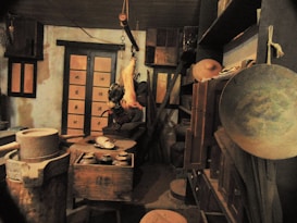 A dimly lit traditional kitchen with various utensils and tools. There's a grinding stone on the left, a hanging meat hook with a cut piece of meat, a wooden table with pots and plates, and shelves filled with different containers and baskets. The room has wooden panels and Japanese-style sliding doors with Japanese characters on them.
