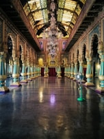 A grand, ornate hall with an elaborate chandelier hanging from the intricately designed ceiling. The hall features rows of detailed columns with golden and turquoise accents leading toward a large, rich red curtain at the far end. The reflective floor adds to the opulence of the space, and a fan can be seen on the right side.
