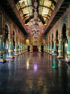 A grand, ornate hall with an elaborate chandelier hanging from the intricately designed ceiling. The hall features rows of detailed columns with golden and turquoise accents leading toward a large, rich red curtain at the far end. The reflective floor adds to the opulence of the space, and a fan can be seen on the right side.