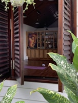 An open wooden window reveals the interior of a serene room with wooden flooring and a high ceiling. Inside, there is a purple yoga mat on the floor and a wall-mounted bookshelf filled with various items such as books and decor. A prominent, serene face painting adorns the bookshelf. The window is flanked by lush green plants with variegated leaves, and a hanging plant is visible at the top.