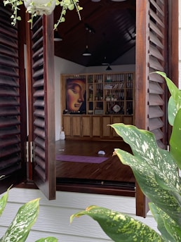 An open wooden window reveals the interior of a serene room with wooden flooring and a high ceiling. Inside, there is a purple yoga mat on the floor and a wall-mounted bookshelf filled with various items such as books and decor. A prominent, serene face painting adorns the bookshelf. The window is flanked by lush green plants with variegated leaves, and a hanging plant is visible at the top.