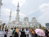 A large white mosque with multiple domes and minarets stands prominently against a partly cloudy sky. In front of the mosque, a diverse group of people are gathered, some wearing traditional attire, and one person is holding a pink umbrella.