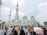 A large white mosque with multiple domes and minarets stands prominently against a partly cloudy sky. In front of the mosque, a diverse group of people are gathered, some wearing traditional attire, and one person is holding a pink umbrella.