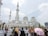 A large white mosque with multiple domes and minarets stands prominently against a partly cloudy sky. In front of the mosque, a diverse group of people are gathered, some wearing traditional attire, and one person is holding a pink umbrella.