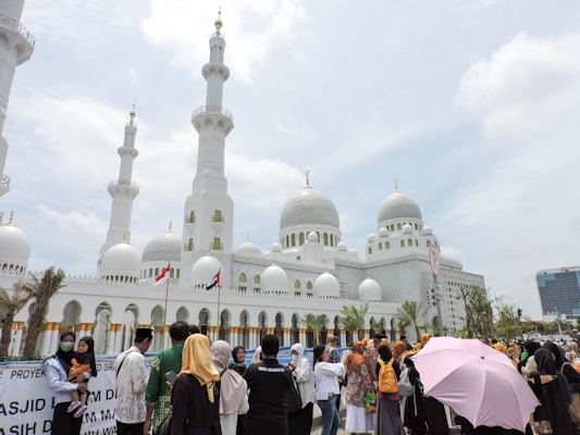 A large white mosque with multiple domes and minarets stands prominently against a partly cloudy sky. In front of the mosque, a diverse group of people are gathered, some wearing traditional attire, and one person is holding a pink umbrella.